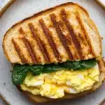 Egg sandwich panini - the image shows a sandwich on a plate. the sandwich is made with two slices of toasted bread, with a golden brown crust on top. the bread appears to be toasted and has a crispy texture. on top of the bread, there is a generous amount of scrambled eggs, which are yellow and runny. there are also a few fresh spinach leaves scattered on top, adding a pop of green color to the dish. the plate is a light beige color with a speckled pattern.