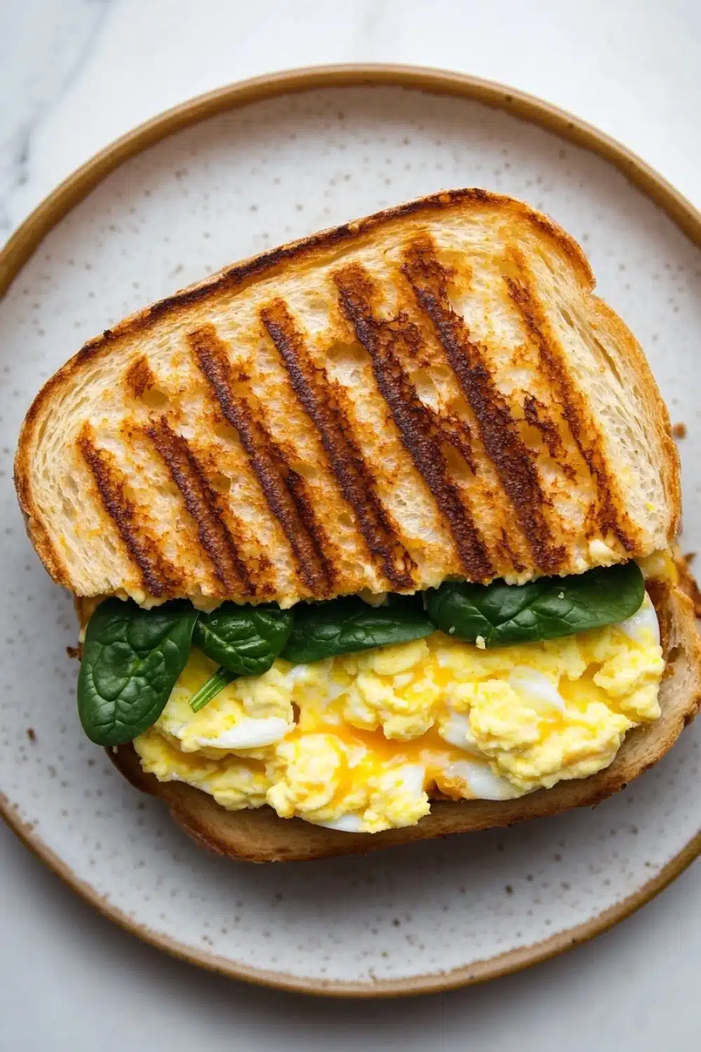 Egg sandwich panini - the image shows a sandwich on a plate. the sandwich is made with two slices of toasted bread, with a golden brown crust on top. the bread appears to be toasted and has a crispy texture. on top of the bread, there is a generous amount of scrambled eggs, which are yellow and runny. there are also a few fresh spinach leaves scattered on top, adding a pop of green color to the dish. the plate is a light beige color with a speckled pattern.