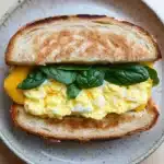Egg sandwich panini - the image shows a sandwich on a plate. the sandwich is made with two slices of toasted bread, with a golden brown crust. on top of the bread, there is a generous amount of scrambled eggs, which are yellow and runny. the eggs are arranged in a circular pattern, with some of them overlapping each other. there are also a few fresh basil leaves scattered on top. the plate is a light beige color with a speckled pattern. the background is blurred, but it appears to be a wooden table.