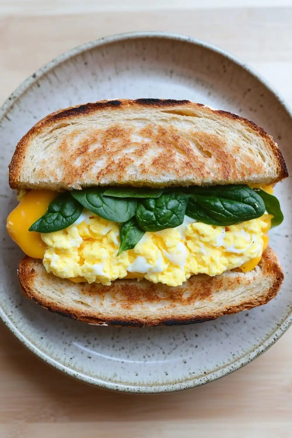 Egg sandwich panini - the image shows a sandwich on a plate. the sandwich is made with two slices of toasted bread, with a golden brown crust. on top of the bread, there is a generous amount of scrambled eggs, which are yellow and runny. the eggs are arranged in a circular pattern, with some of them overlapping each other. there are also a few fresh basil leaves scattered on top. the plate is a light beige color with a speckled pattern. the background is blurred, but it appears to be a wooden table.