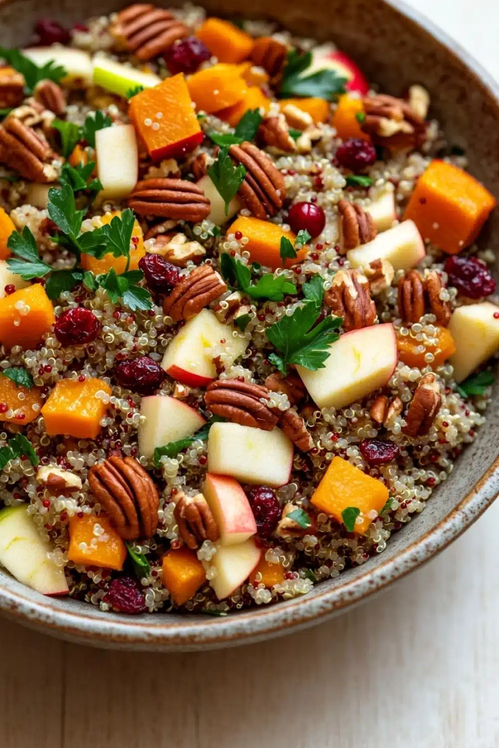 Fall harvest quinoa salad - the image is a close-up of a bowl of quinoa salad. the bowl is made of ceramic and is placed on a wooden table. the quinoa is a light brown color and is mixed with various ingredients such as sliced apples, butternut squash, cranberries, and pecans. the salad is garnished with fresh parsley. the colors of the ingredients are vibrant and the dish looks fresh and appetizing.