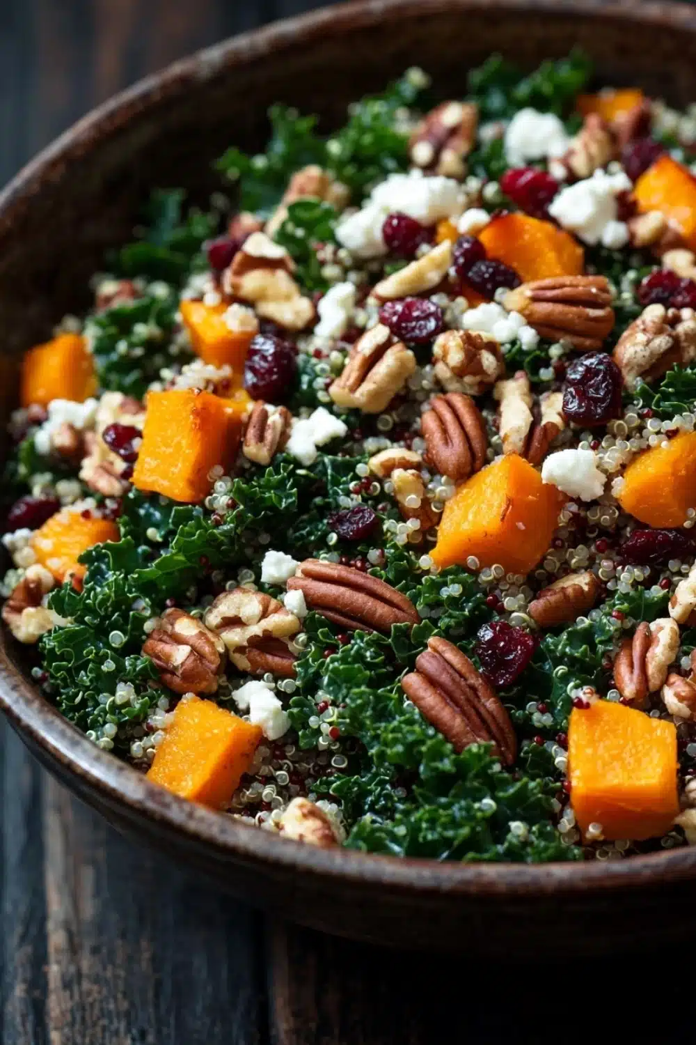 Fall harvest quinoa salad - the image is a close-up of a bowl of salad. the bowl is made of dark brown ceramic and is placed on a wooden table. the salad is made up of kale, butternut squash, cranberries, and pecans, and is topped with crumbled feta cheese. the colors of the ingredients are vibrant and the dish looks fresh and appetizing.