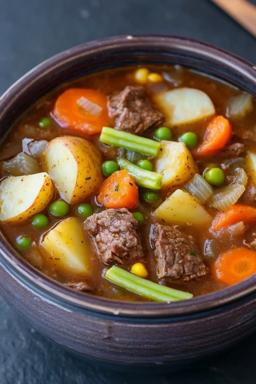 Freezer vegetable beef stew - the image is a close-up of a bowl of beef stew. the bowl is made of dark brown ceramic and is sitting on a dark grey countertop. the stew is made up of chunks of beef, potatoes, carrots, green peas, and onions. the beef appears to be cooked medium-rare and is visible in the broth. the vegetables are bright orange and look fresh and vibrant. the broth is thick and thick, and the vegetables are arranged in a way that creates a colorful and appetizing appearance.
