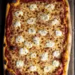 Full sheet pan pizza - the image is of a freshly baked pizza on a wooden cutting board. the pizza is rectangular in shape and has a golden brown crust. it is topped with melted cheese and small black pepper sprinkled on top. the cheese appears to be melted and bubbly, and there are small pieces of tomato sauce scattered throughout the pizza. the background is a dark wooden table with a rustic texture. the overall mood of the image is warm and inviting.