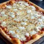 Full sheet pan pizza - the image shows a freshly baked pizza on a wooden cutting board. the pizza has a golden brown crust and is topped with melted cheese and small chunks of tomato sauce. the cheese is white and appears to be melted and bubbly. there are also small black pepper sprinkled on top of the cheese. in the background, there is a white pitcher and a gray cloth napkin. the cutting board is placed on a dark countertop.