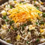 Garlic butter beef rice skillet - the image is a close-up of a bowl of food. the bowl is made of ceramic and is sitting on a white marble countertop. the food appears to be a dish made with rice, ground beef, and cheese. the rice is white and fluffy, and the ground beef is a dark brown color. the cheese is a bright yellow color and is scattered on top of the rice. there are also small pieces of diced potatoes and chopped parsley scattered throughout the dish. the dish looks freshly cooked and ready to eat.
