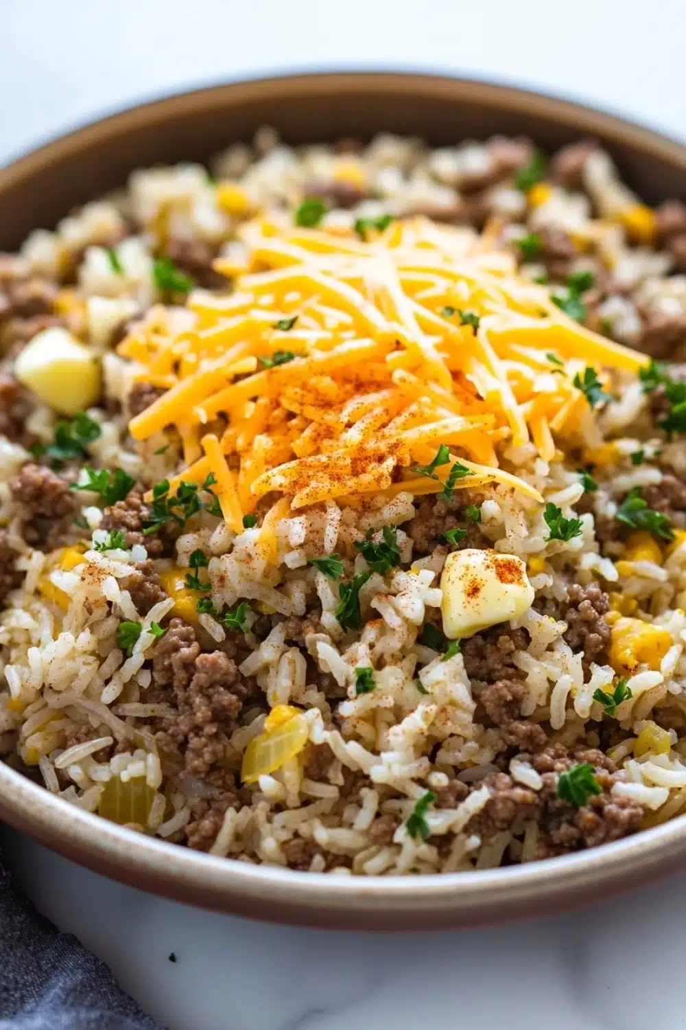 Garlic butter beef rice skillet - the image is a close-up of a bowl of food. the bowl is made of ceramic and is sitting on a white marble countertop. the food appears to be a dish made with rice, ground beef, and cheese. the rice is white and fluffy, and the ground beef is a dark brown color. the cheese is a bright yellow color and is scattered on top of the rice. there are also small pieces of diced potatoes and chopped parsley scattered throughout the dish. the dish looks freshly cooked and ready to eat.