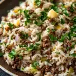 Garlic butter beef rice skillet - the image is a close-up of a dish in a brown bowl. the dish appears to be a type of rice or risotto, with chunks of ground beef and diced potatoes scattered throughout. the rice is white and fluffy, and the ground beef is a dark brown color. there are also a few sprigs of fresh parsley scattered throughout the dish, adding a pop of green color to the dish. the bowl is sitting on a dark grey countertop.