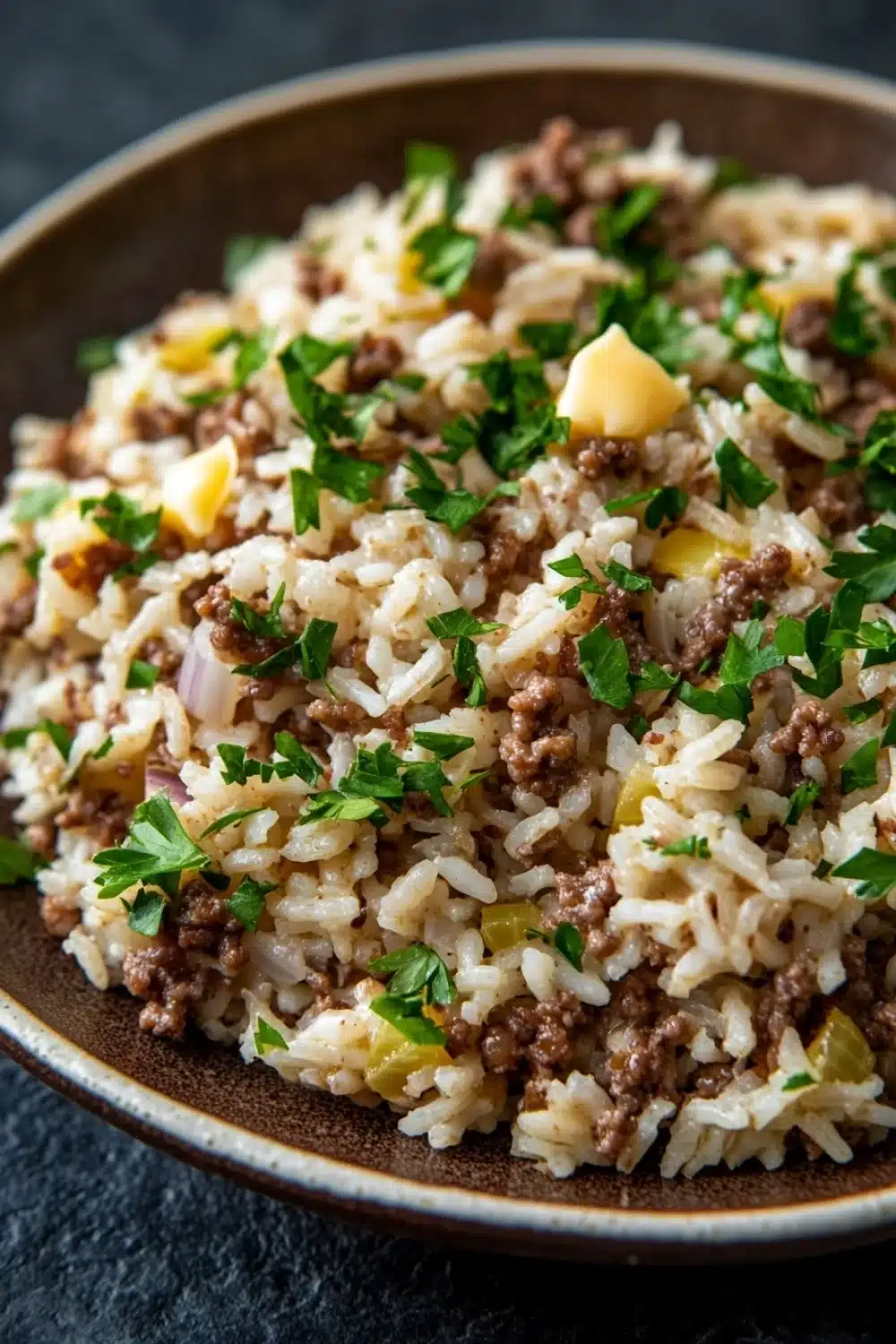 Garlic butter beef rice skillet - the image is a close-up of a dish in a brown bowl. the dish appears to be a type of rice or risotto, with chunks of ground beef and diced potatoes scattered throughout. the rice is white and fluffy, and the ground beef is a dark brown color. there are also a few sprigs of fresh parsley scattered throughout the dish, adding a pop of green color to the dish. the bowl is sitting on a dark grey countertop.
