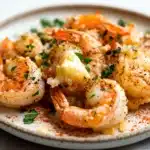 Garlic butter shrimp bake - the image shows a plate of cooked shrimp on a white plate. the shrimp are orange in color and appear to be seasoned with herbs and spices. they are arranged in a circular pattern on the plate, with some overlapping each other. on top of the shrimp, there is a small piece of butter, which is placed on top. the plate is garnished with a sprig of parsley. the background is blurred, but it appears to be a kitchen countertop.