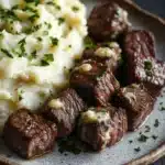 Garlic butter steak bites and mash - the image shows a plate of steak and mashed potatoes. the steak is cooked medium-rare and has a golden brown crust on the outside. the mashed potatoes are creamy and fluffy, and they are topped with a dollop of white sauce and garnished with chopped parsley. the plate is made of dark wood and is placed on a wooden table. the background is blurred, but it appears to be a rustic kitchen countertop.