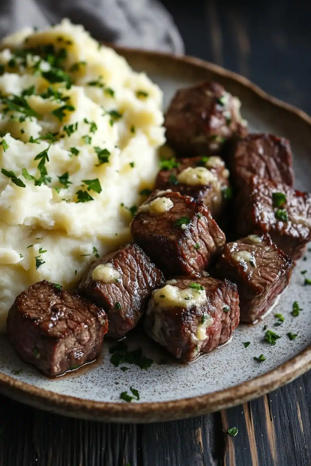 Garlic butter steak bites and mash - the image shows a plate of steak and mashed potatoes. the steak is cooked medium-rare and has a golden brown crust on the outside. the mashed potatoes are creamy and fluffy, and they are topped with a dollop of white sauce and garnished with chopped parsley. the plate is made of dark wood and is placed on a wooden table. the background is blurred, but it appears to be a rustic kitchen countertop.