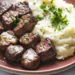 Garlic butter steak bites and mash - the image shows a plate of food with a serving of mashed potatoes and meatballs. the mashed potatoes are creamy and white, and the meatballs are dark brown in color. the plate is made of a light-colored ceramic or porcelain material and is garnished with fresh parsley. the meatballs appear to be cooked and have a crispy texture. the dish looks appetizing and ready to eat.