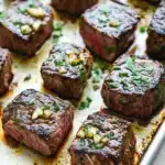 Garlic butter steak bites chuck roast - the image shows a baking tray lined with parchment paper. on the tray, there are several pieces of cooked steak arranged in a single layer. the steak appears to be medium-rare and has a pinkish-red color. it is garnished with chopped parsley and chopped nuts. the tray is sitting on a white countertop.