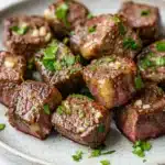 Garlic butter steak bites ingredients - the image shows a plate of cooked steak cubes. the steak cubes are arranged in a circular pattern on a white plate with a speckled texture. they are covered in a dark brown sauce and garnished with chopped parsley. the sauce appears to be drizzled over the top of the steak, giving it a glossy sheen. the dish looks appetizing and ready to eat.