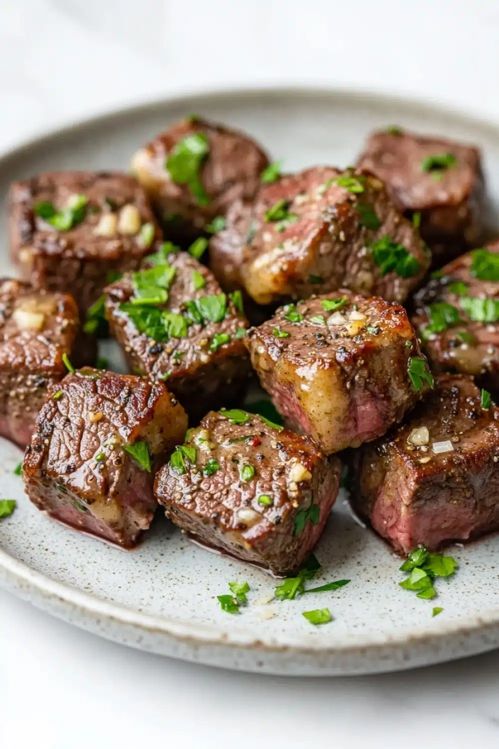 Garlic butter steak bites ingredients - the image shows a plate of cooked steak cubes. the steak cubes are arranged in a circular pattern on a white plate with a speckled texture. they are covered in a dark brown sauce and garnished with chopped parsley. the sauce appears to be drizzled over the top of the steak, giving it a glossy sheen. the dish looks appetizing and ready to eat.