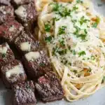 Garlic butter steak bites with linguine - the image shows a plate of food with a serving of spaghetti and meat. the plate is made of light-colored ceramic and has a speckled texture. on the left side of the plate, there are several pieces of grilled steak arranged in a circular pattern. the steak appears to be cooked medium-rare and is garnished with a sprinkle of parmesan cheese and fresh herbs. next to the steak, there is a pile of spaghetti with a generous amount of grated cheese on top. the dish is served on a wooden table with a fork and knife on the side.