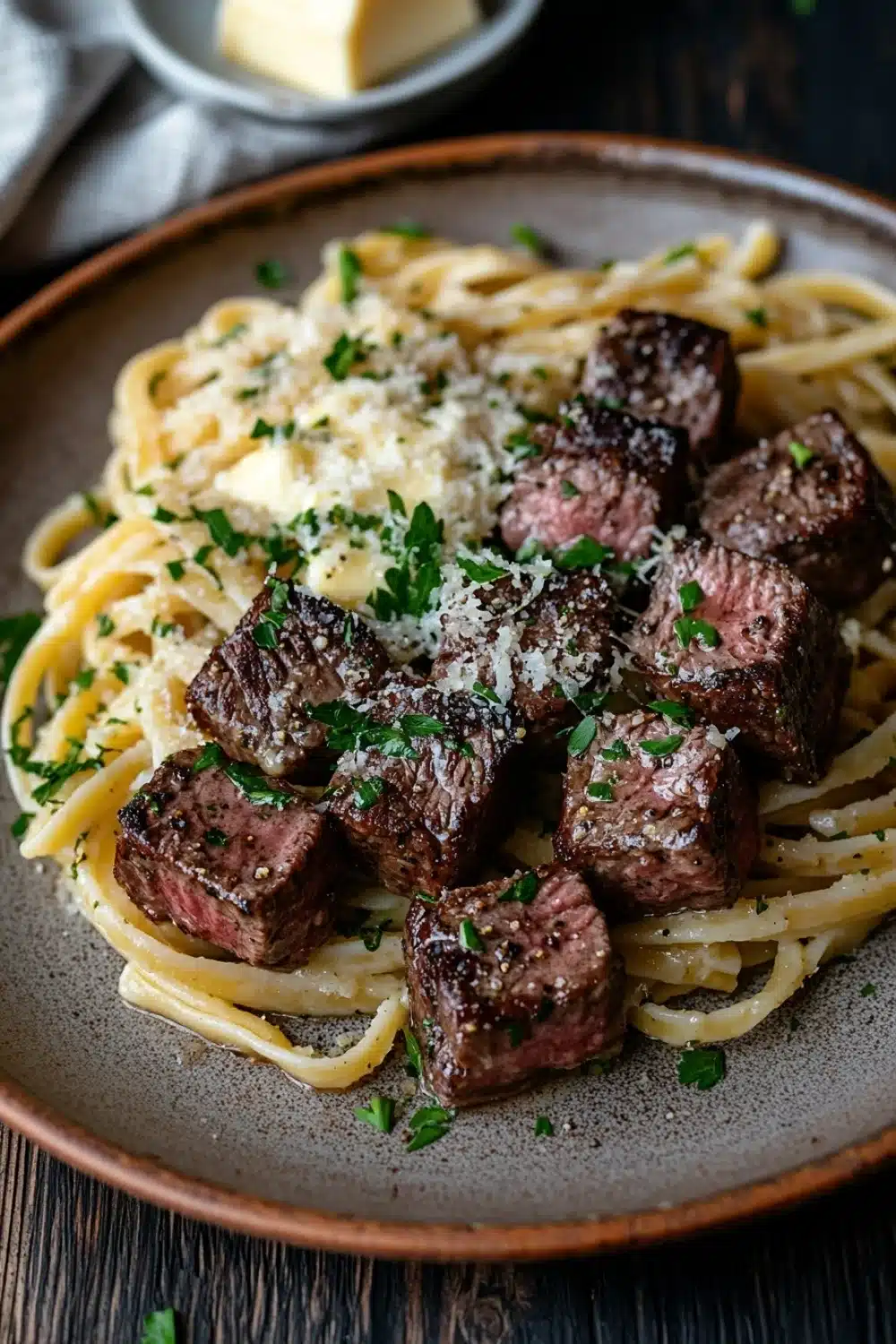 Garlic butter steak bites with linguine - the image shows a plate of spaghetti with chunks of beef and parmesan cheese on top. the plate is made of brown ceramic and is placed on a wooden table with a white napkin and a small bowl of butter in the background. the spaghetti is cooked al dente and is topped with a sprinkle of chopped parsley. the beef appears to be cooked medium-rare and has a dark brown color. the cheese is melted and bubbly on top, and there is a sprinkle on top of the meat. the overall presentation of the dish is elegant and appetizing.