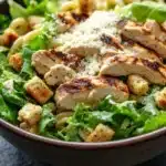 Gluten free chicken caesar pasta salad - the image is a close-up of a bowl of pasta salad. the bowl is made of dark brown ceramic and is placed on a dark grey countertop. the salad is made up of a bed of green leafy vegetables, possibly lettuce or arugula, and is topped with sliced chicken and croutons. the chicken is cooked to a golden brown color and appears to be seasoned with herbs and spices. there is also a sprinkle of parmesan cheese on top of the chicken. in the background, there is a small bowl of creamy dressing.