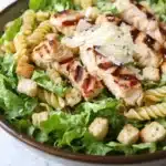 Gluten free chicken caesar pasta salad - the image is a close-up of a bowl of pasta salad. the salad is made up of spiral pasta, arugula, and croutons. the pasta is cooked al dente and appears to be seasoned with herbs and spices. on top of the pasta, there are grilled chicken pieces that have been grilled to a golden brown color. the chicken pieces are arranged in a circular pattern and are scattered throughout the salad. there is also a sprinkle of grated parmesan cheese on top. the bowl is sitting on a white marble countertop.