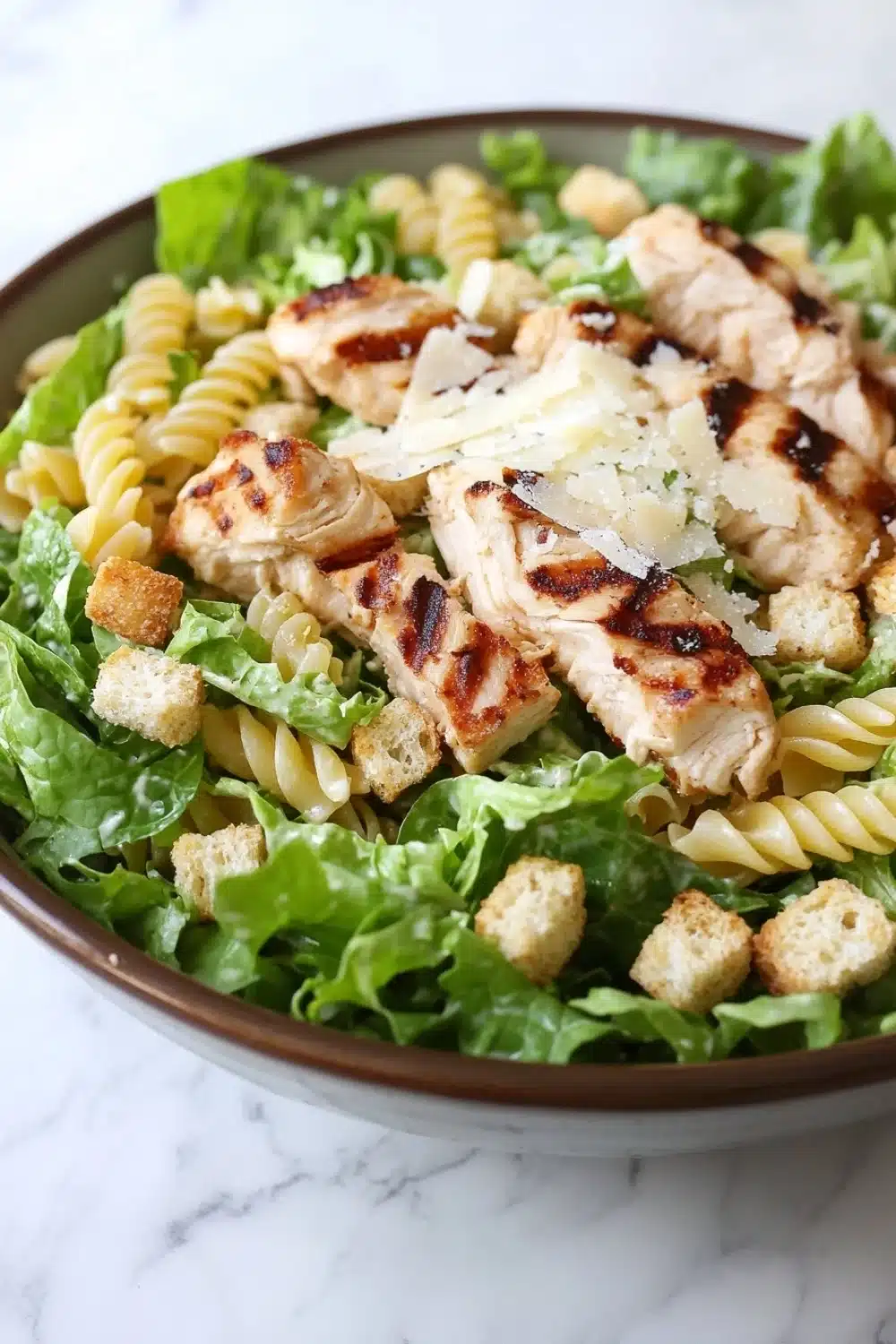 Gluten free chicken caesar pasta salad - the image is a close-up of a bowl of pasta salad. the salad is made up of spiral pasta, arugula, and croutons. the pasta is cooked al dente and appears to be seasoned with herbs and spices. on top of the pasta, there are grilled chicken pieces that have been grilled to a golden brown color. the chicken pieces are arranged in a circular pattern and are scattered throughout the salad. there is also a sprinkle of grated parmesan cheese on top. the bowl is sitting on a white marble countertop.