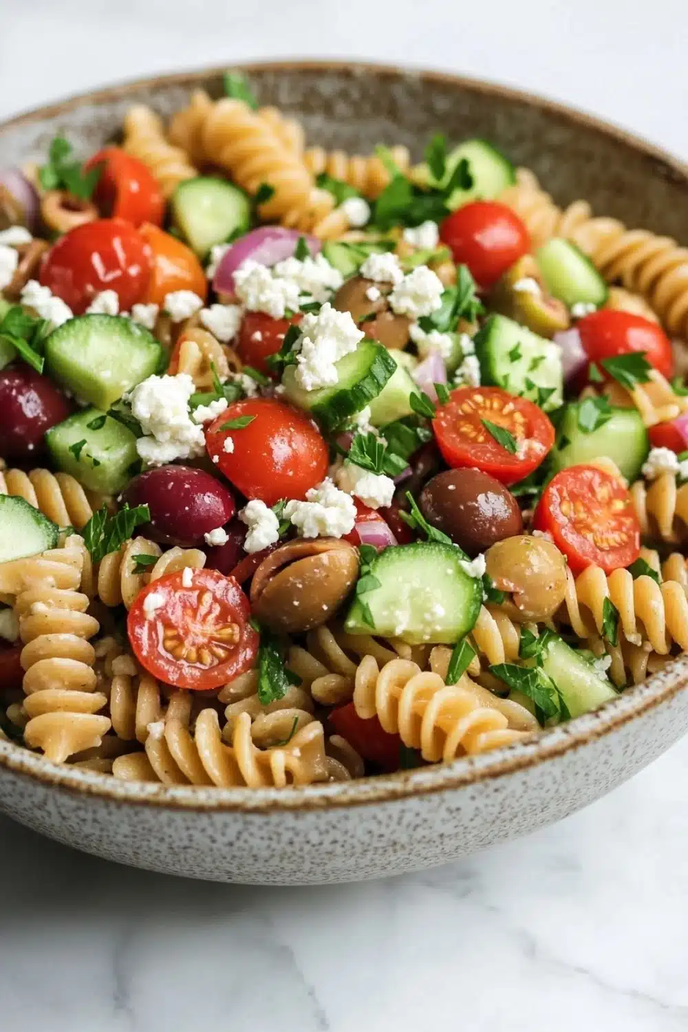 Greek pasta salad vegan - the image is of a bowl of pasta salad. the bowl is made of ceramic and is sitting on a white marble countertop. the pasta salad is made up of spiral pasta, cherry tomatoes, cucumbers, olives, and feta cheese. the colors of the ingredients are vibrant and the dish looks fresh and appetizing.