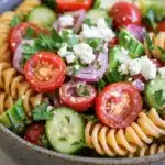 Greek pasta salad vegan - the image is a close-up of a bowl of pasta salad. the bowl is made of ceramic and is sitting on a white countertop. the pasta salad is made up of spiral pasta, sliced tomatoes, cucumbers, red onions, and feta cheese. the tomatoes are bright red and appear to be ripe, while the cucumbers are green and juicy. the feta is crumbled and scattered throughout the salad, adding a creamy texture. there are also some fresh herbs and cilantro scattered throughout. the overall color scheme of the salad is vibrant and appetizing.