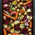 Greek roasted root vegetables - the image shows a rectangular baking tray filled with roasted vegetables. the vegetables are arranged in a single layer on the tray, with some overlapping each other. the carrots, beets, and onions are cut into small cubes and are garnished with chopped parsley. the tray is sitting on a dark grey countertop. the overall appearance of the dish is colorful and appetizing.