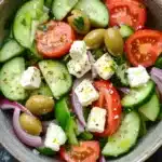 Greek salad - the image is a close-up of a colorful salad in a bowl. the salad is made up of sliced cucumbers, cherry tomatoes, olives, red onions, and feta cheese. the cucumbers are cut into thin slices and are arranged in a circular pattern. the tomatoes are bright red and appear to be ripe and juicy. the olives are scattered throughout the salad, adding a pop of green color to the dish. the feta is crumbled and adds a creamy texture to the overall appearance. the bowl is made of light-colored wood and is sitting on a dark grey countertop.