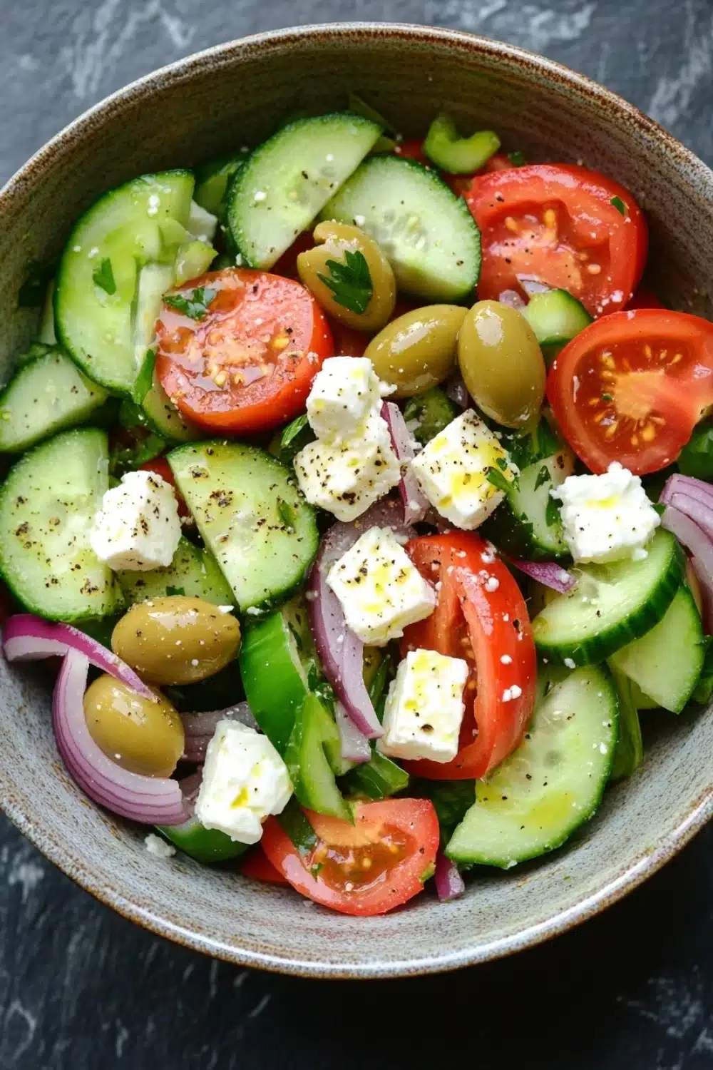 Greek salad - the image is a close-up of a colorful salad in a bowl. the salad is made up of sliced cucumbers, cherry tomatoes, olives, red onions, and feta cheese. the cucumbers are cut into thin slices and are arranged in a circular pattern. the tomatoes are bright red and appear to be ripe and juicy. the olives are scattered throughout the salad, adding a pop of green color to the dish. the feta is crumbled and adds a creamy texture to the overall appearance. the bowl is made of light-colored wood and is sitting on a dark grey countertop.