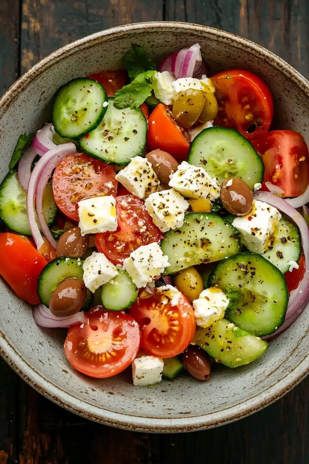 Greek salad - the image is a close-up of a colorful salad in a bowl. the salad is made up of sliced cucumbers, cherry tomatoes, red onions, olives, and feta cheese. the cucumbers are cut into thin slices and are arranged in a circular pattern. the tomatoes are bright red and appear to be ripe and juicy. the olives are scattered throughout the salad, adding a pop of green color to the dish. the feta is crumbled and adds a creamy texture to the overall appearance. the bowl is made of light-colored ceramic and is sitting on a wooden table.