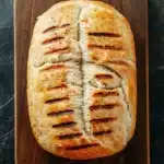Grilled chicken sandwich bread - the image shows a loaf of bread on a wooden cutting board. the bread appears to be freshly baked and has a golden brown crust on top. the slices of bread are arranged in a criss-cross pattern, with each slice slightly overlapping the one below it. the cutting board is placed on a dark grey marble countertop. the overall mood of the image is rustic and appetizing.