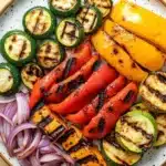 Grilled vegetables in air fryer - the image is a close-up of a plate of grilled vegetables. the plate is white with a speckled pattern and is placed on a wooden table. the vegetables are arranged in a circular pattern on the plate. on the left side of the plate, there are several zucchini and bell peppers, which are cut into small cubes and have a golden-brown color. next to the peppers, there is a pile of sliced red onions. the peppers are grilled to a golden brown color and appear to be seasoned with herbs and spices. the overall appearance of the dish is colorful and appetizing.