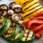 Grilled vegetables in air fryer - the image is a close-up of a plate of grilled vegetables. the plate is made of a light-colored ceramic with a speckled pattern. on the left side of the plate, there are several slices of zucchini, which are grilled to a golden brown color and have a crispy texture. next to them, there is a pile of sliced yellow and red bell peppers and sliced mushrooms. the vegetables are arranged in a circular pattern on the plate. the background is dark, making the colors of the vegetables stand out.