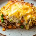Ground beef casserole with hashbrowns - the image shows a plate of lasagna on a white tablecloth. the lasagna is layered with layers of ground beef, peas, carrots, and other vegetables. the top layer is covered in melted cheese, which is a bright yellow color. the cheese appears to be melted and bubbly, and the edges of the lasagna are golden brown. the plate is round and has a speckled pattern around the edges.
