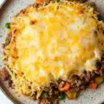 Ground beef casserole with hashbrowns - the image is a close-up of a dish on a white marble countertop. the dish appears to be a casserole or lasagna, with a layer of ground beef and melted cheese on top. the cheese is golden brown and bubbly, and it is spread evenly over the top of the dish. there are also small pieces of green peas scattered throughout the dish, adding a pop of color. the plate is round and has a speckled pattern around the edges.