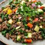 Ground beef dinner high protein - the image is a close-up of a plate of food. the plate is round and has a light brown color. the food appears to be a salad with a variety of ingredients, including quinoa, sliced cucumbers, red onions, yellow bell peppers, and green leafy vegetables. the quinoa is mixed with herbs and spices, and the vegetables are arranged in a colorful and appetizing manner. the dish is garnished with fresh cilantro leaves. the background is a dark grey countertop.