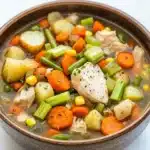 Hearty chicken vegetable soup crock pot - the image is a close-up of a bowl of soup. the soup appears to be a vegetable soup with chunks of chicken, carrots, celery, corn, and potatoes. the vegetables are mixed in with the soup, creating a colorful and appetizing appearance. the bowl is made of ceramic and has a dark brown color. the background is a white marble countertop.