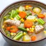 Hearty chicken vegetable soup crock pot - the image is a close-up of a bowl of soup. the soup appears to be a vegetable soup with chunks of chicken, carrots, celery, corn, and other vegetables mixed in. the bowl is made of ceramic and has a brown and white speckled pattern. the broth is a light brown color and looks thick and creamy. the vegetables are bright orange and appear to be fresh and vibrant. the chicken is visible in the center of the soup, with a sprinkle of herbs on top. the background is white, making the colors of the vegetables stand out.