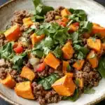 High protein beef and sweet potato bowl - the image is a close-up of a plate of food. the plate is made of a light-colored ceramic bowl with a beige rim. the food appears to be a salad with a variety of ingredients, including quinoa, sweet potatoes, spinach, and red bell peppers. the quinoa is a dark brown color and is mixed with the sweet potatoes and spinach. the dish is garnished with fresh cilantro leaves. the background is dark, making the colors of the food stand out.