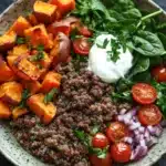 High protein beef and sweet potato bowl - the image is a close-up of a bowl of food. the bowl is made of a light-colored ceramic with a speckled pattern. on the left side of the bowl, there are sliced sweet potatoes arranged in a circular pattern. next to the sweet potatoes, there is a bed of quinoa, sliced red onions, cherry tomatoes, and a dollop of sour cream. the quinoa is a dark brown color and appears to be seasoned with herbs and spices. the tomatoes are bright red and appear to be ripe and juicy. the spinach leaves are a vibrant green color and are scattered throughout the bowl. the overall presentation of the dish is colorful and appetizing.