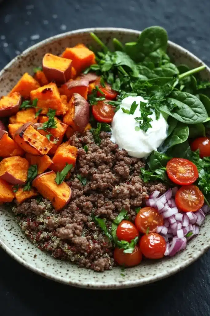 High protein beef and sweet potato bowl - the image is a close-up of a bowl of food. the bowl is made of a light-colored ceramic with a speckled pattern. on the left side of the bowl, there are sliced sweet potatoes arranged in a circular pattern. next to the sweet potatoes, there is a bed of quinoa, sliced red onions, cherry tomatoes, and a dollop of sour cream. the quinoa is a dark brown color and appears to be seasoned with herbs and spices. the tomatoes are bright red and appear to be ripe and juicy. the spinach leaves are a vibrant green color and are scattered throughout the bowl. the overall presentation of the dish is colorful and appetizing.