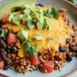 High protein beef enchilada bowls - the image is a close-up of a plate of food. the plate is white with a blue rim and is placed on a wooden table. the food appears to be a dish made with ground beef, black beans, and rice. on top of the beef, there is a layer of melted cheese and sliced avocado. there are also some fresh cilantro scattered around the plate. the background is blurred, but it seems like the plate is in a kitchen or dining area.