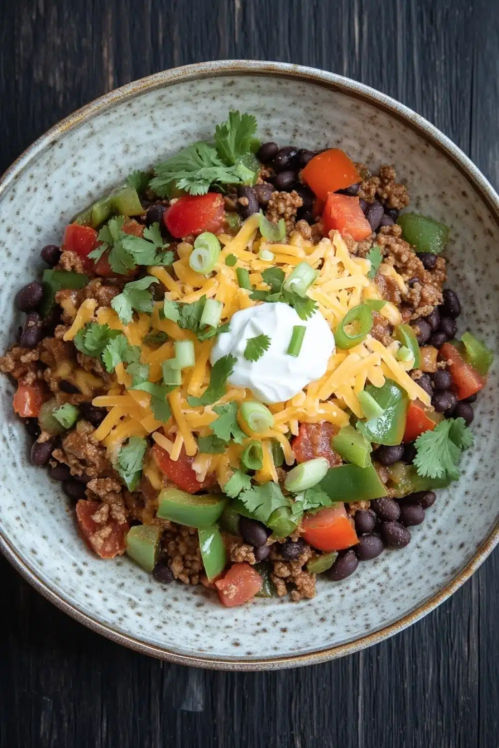 High protein beef enchilada bowls - the image is a close-up of a bowl of chili. the bowl is made of ceramic and has a speckled pattern. the chili is made up of black beans, ground beef, diced tomatoes, green onions, and shredded cheese. there is also a dollop of sour cream on top of the chili. a sprig of cilantro is scattered throughout the dish. the background is a dark wooden table.
