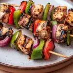 High protein chicken kebab - the image shows a plate of grilled chicken skewers on a wooden table. the skewers are made up of small pieces of chicken, red and green bell peppers, and red onions. the chicken appears to be seasoned with herbs and spices, giving it a golden brown color. the vegetables are arranged in a circular pattern on the skewers, with some pieces overlapping each other. the plate is round and has a light brown rim. the background is blurred, but it seems to be a kitchen countertop.