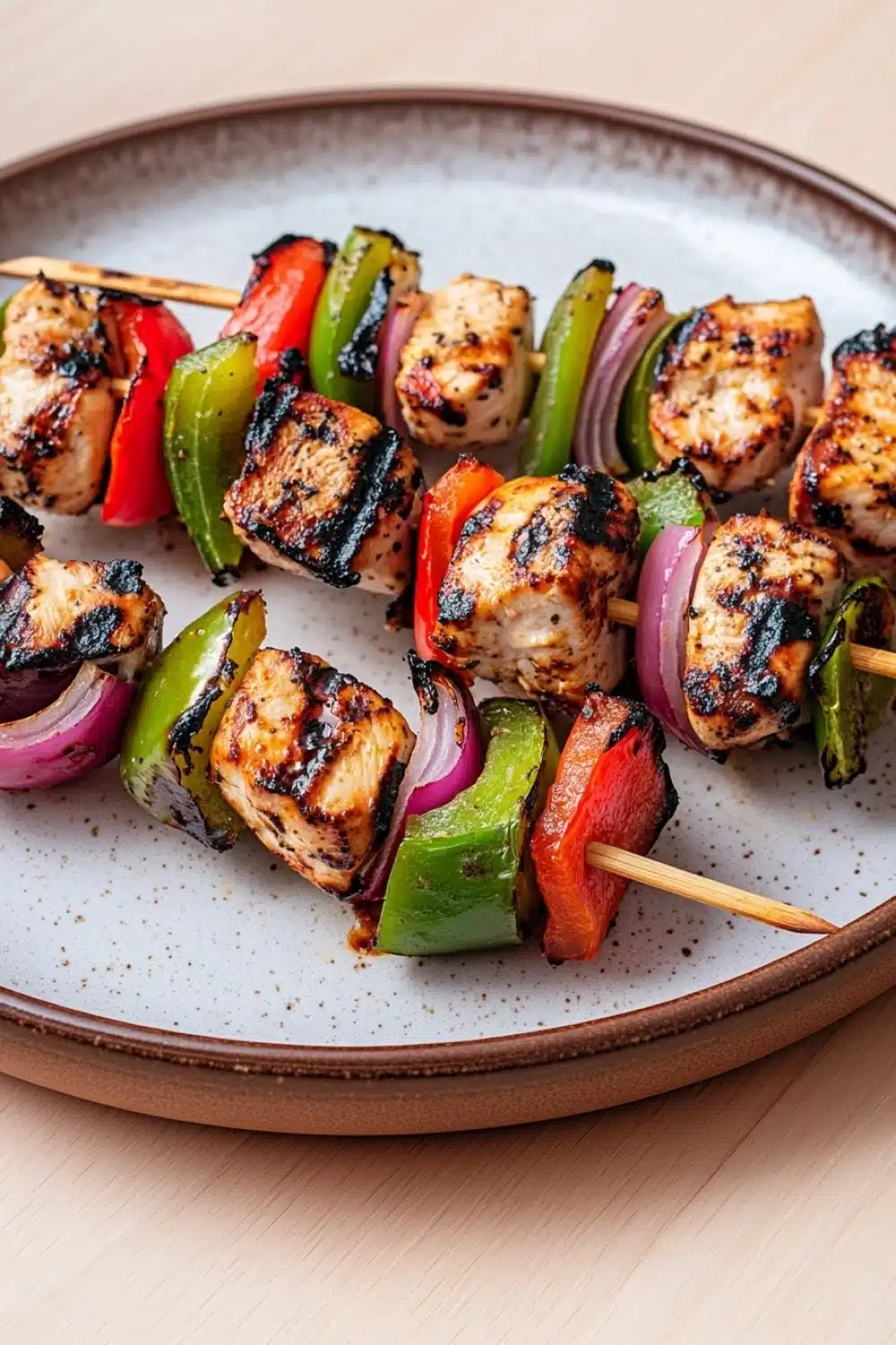High protein chicken kebab - the image shows a plate of grilled chicken skewers on a wooden table. the skewers are made up of small pieces of chicken, red and green bell peppers, and red onions. the chicken appears to be seasoned with herbs and spices, giving it a golden brown color. the vegetables are arranged in a circular pattern on the skewers, with some pieces overlapping each other. the plate is round and has a light brown rim. the background is blurred, but it seems to be a kitchen countertop.