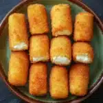 Homemade mozzarella sticks without string cheese - the image shows a plate of nine pieces of breaded fish sticks arranged in a circular pattern on a dark green plate. the fish sticks are golden brown in color and appear to be crispy on the outside and soft on the inside. they are arranged in two rows of three, with the top row slightly overlapping the bottom row. the plate is sitting on a black surface, possibly a table or countertop.