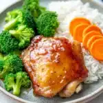 Honey garlic chicken thigh dinner - the image shows a plate of food with a serving of chicken and rice. the chicken is cooked to a golden brown color and appears to be seasoned with sesame seeds. the rice is white and fluffy, and there are a few pieces of broccoli scattered around the plate. on the right side of the plate, there are sliced carrots. the plate is sitting on a white marble countertop.