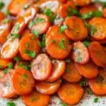 Honey glazed carrots vegan - the image shows a plate of cooked carrots. the carrots are bright orange in color and appear to be seasoned with herbs and spices. they are arranged in a circular pattern on the plate, with some overlapping each other. the plate is white with a speckled pattern and is sitting on a wooden table. the background is blurred, but it appears to be a kitchen countertop.