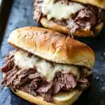 Hot roast beef sandwich slow cooker - the image shows two sandwiches on a black baking tray. the sandwiches are made with a toasted bun and are filled with shredded beef and melted cheese. the beef appears to be cooked medium-rare and the cheese is a creamy white color. the tray is lined with parchment paper and there is a blurred background.